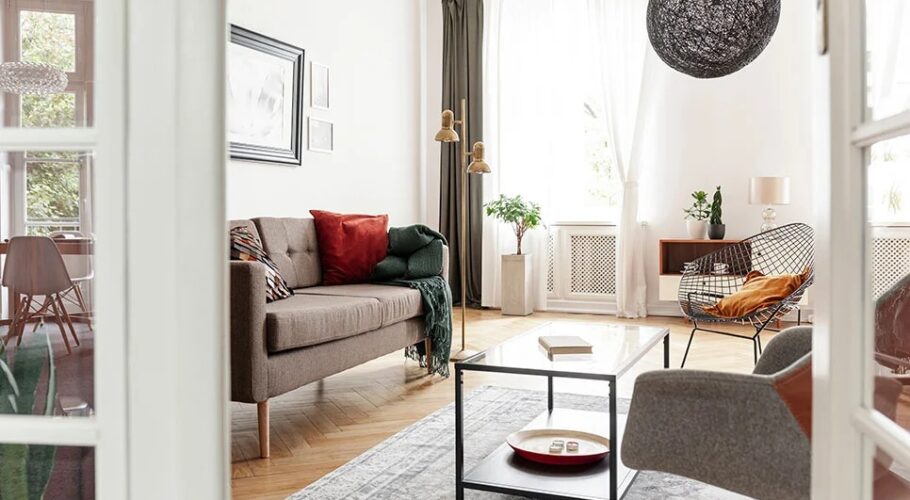 Eklektyczny salon z brązową sofą, stolikiem i fotelem, widziany przez przeszklone drzwi.