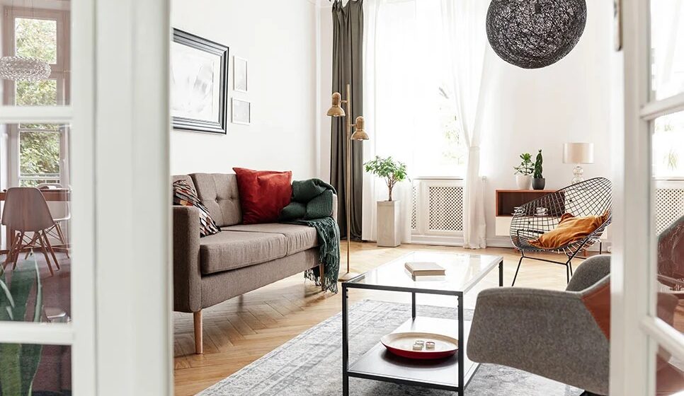 Eklektyczny salon z brązową sofą, stolikiem i fotelem, widziany przez przeszklone drzwi.
